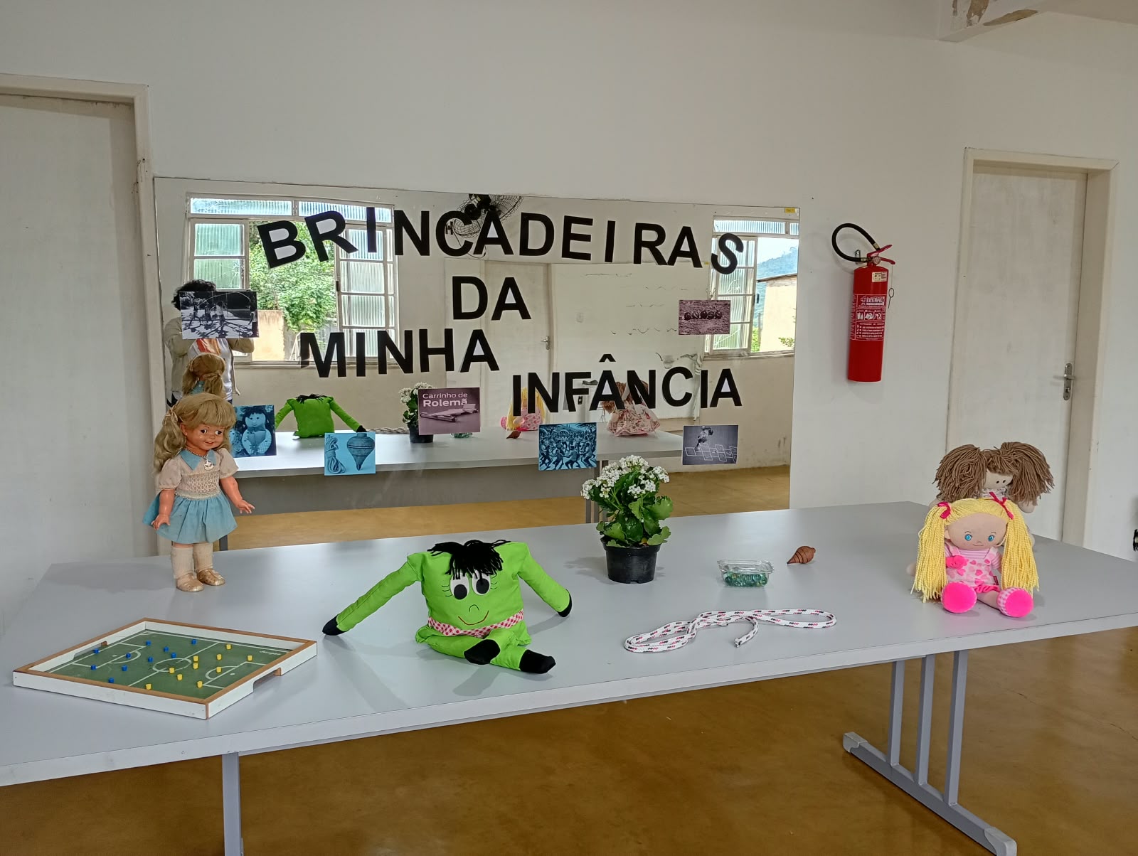 Mesa com bonecas e brinquedos antigos diante do painel Brincadeiras da Minha Infância